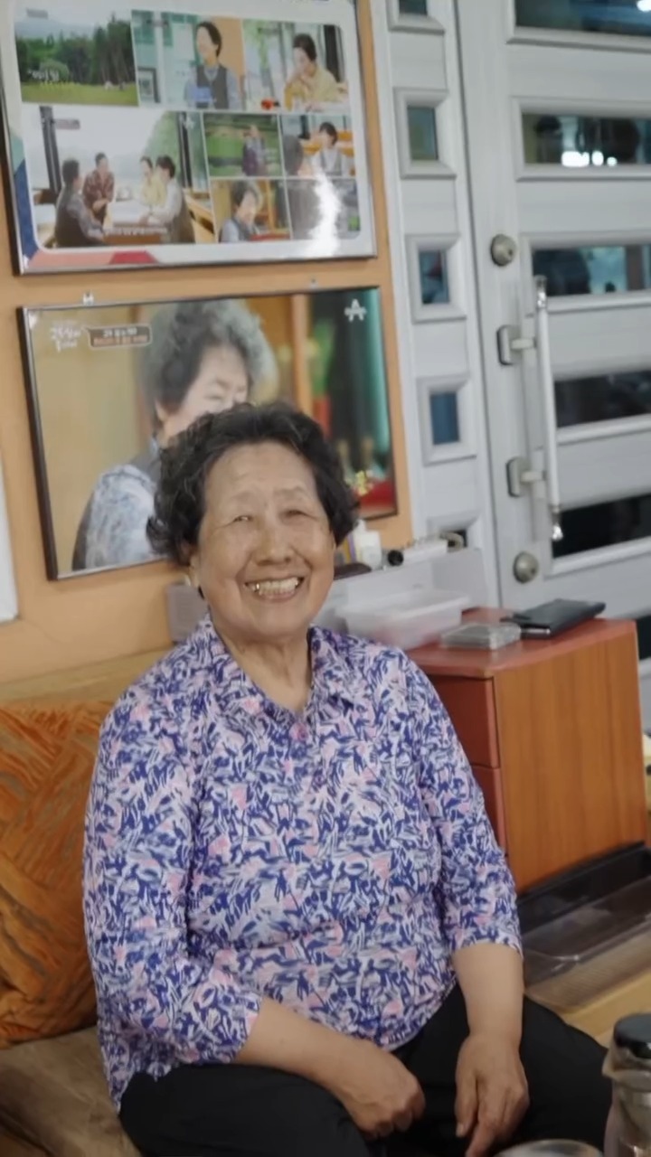 Ahn Hangja sitting in the tea room at Seogwi Dawon, smiling, with framed photographs and press clippings on the wall behind her