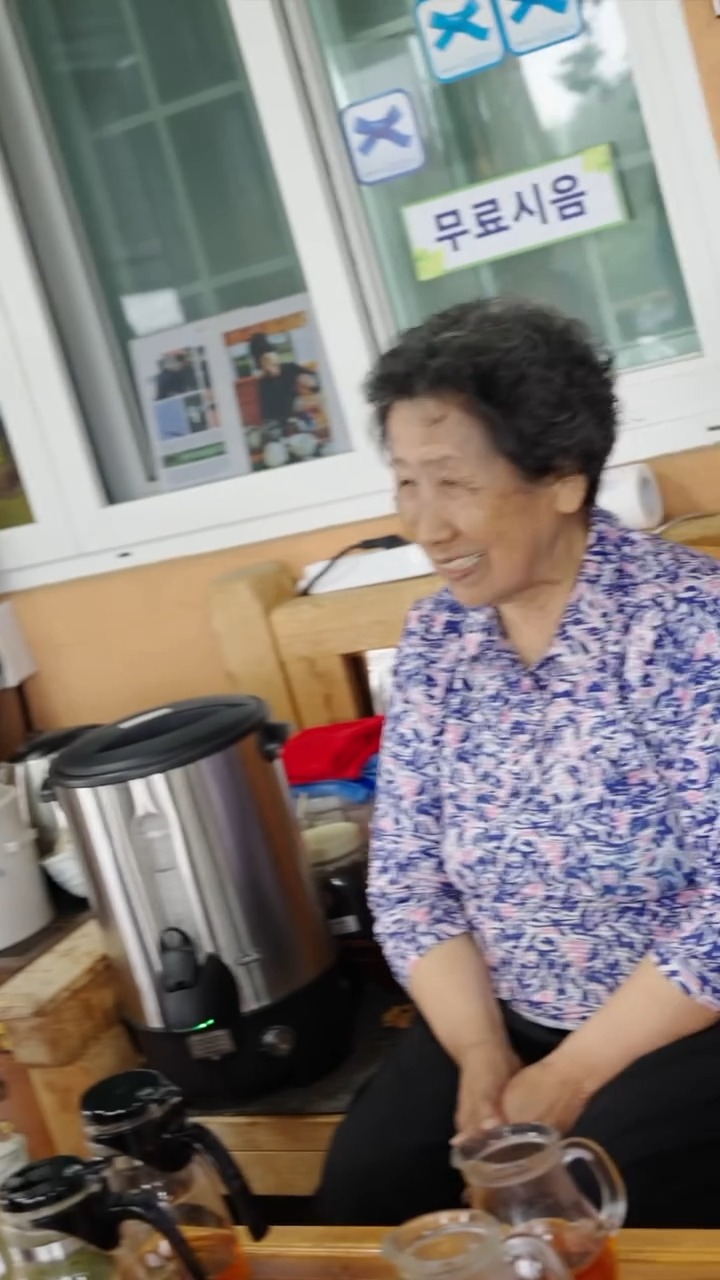 Ahn Hangja beside the hot water urn at Seogwi Dawon, under a hand-written sign offering a free tasting